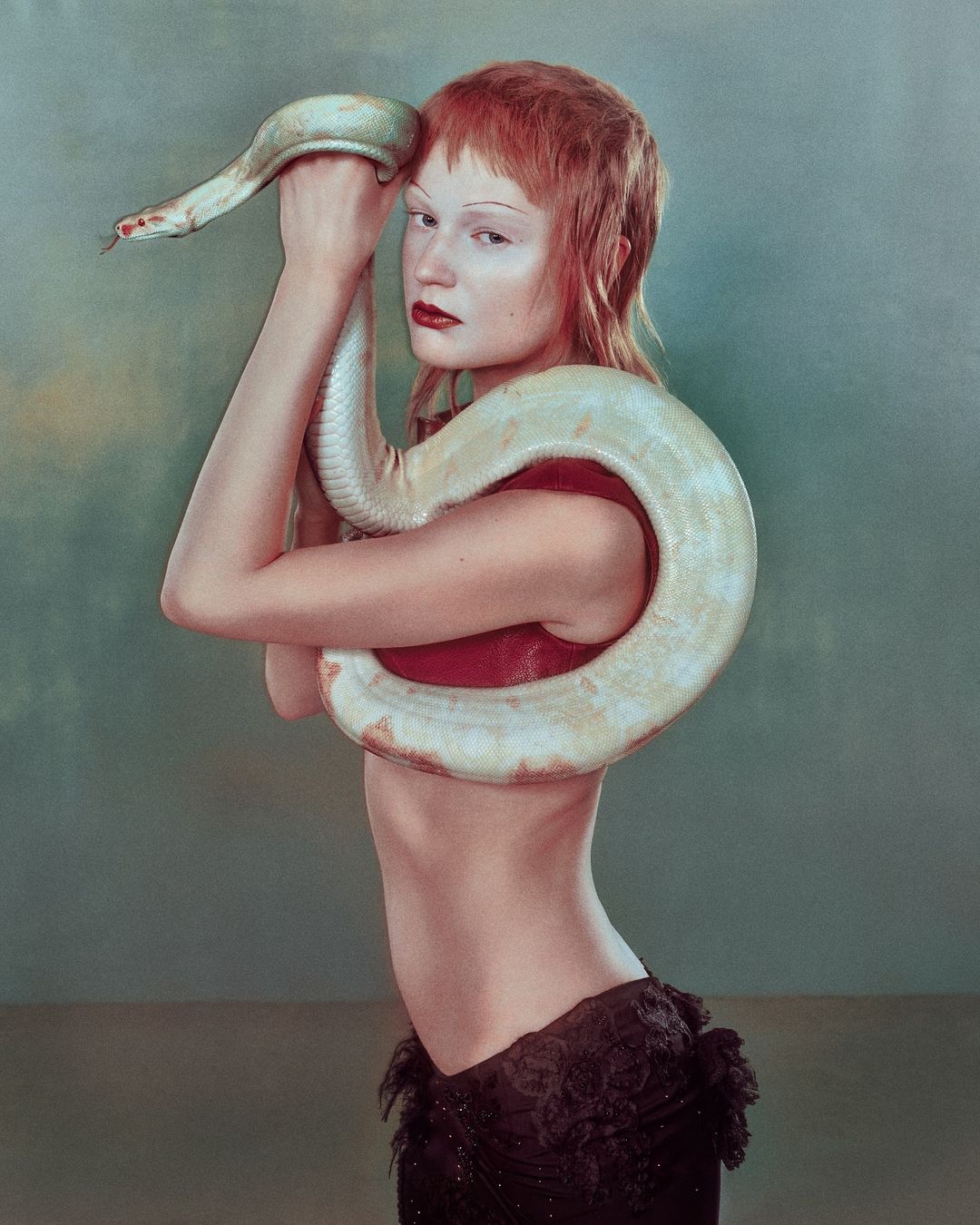 An androgynous model with pale skin and orange hair poses with a large albino snake draped around the shoulders, against a muted background.