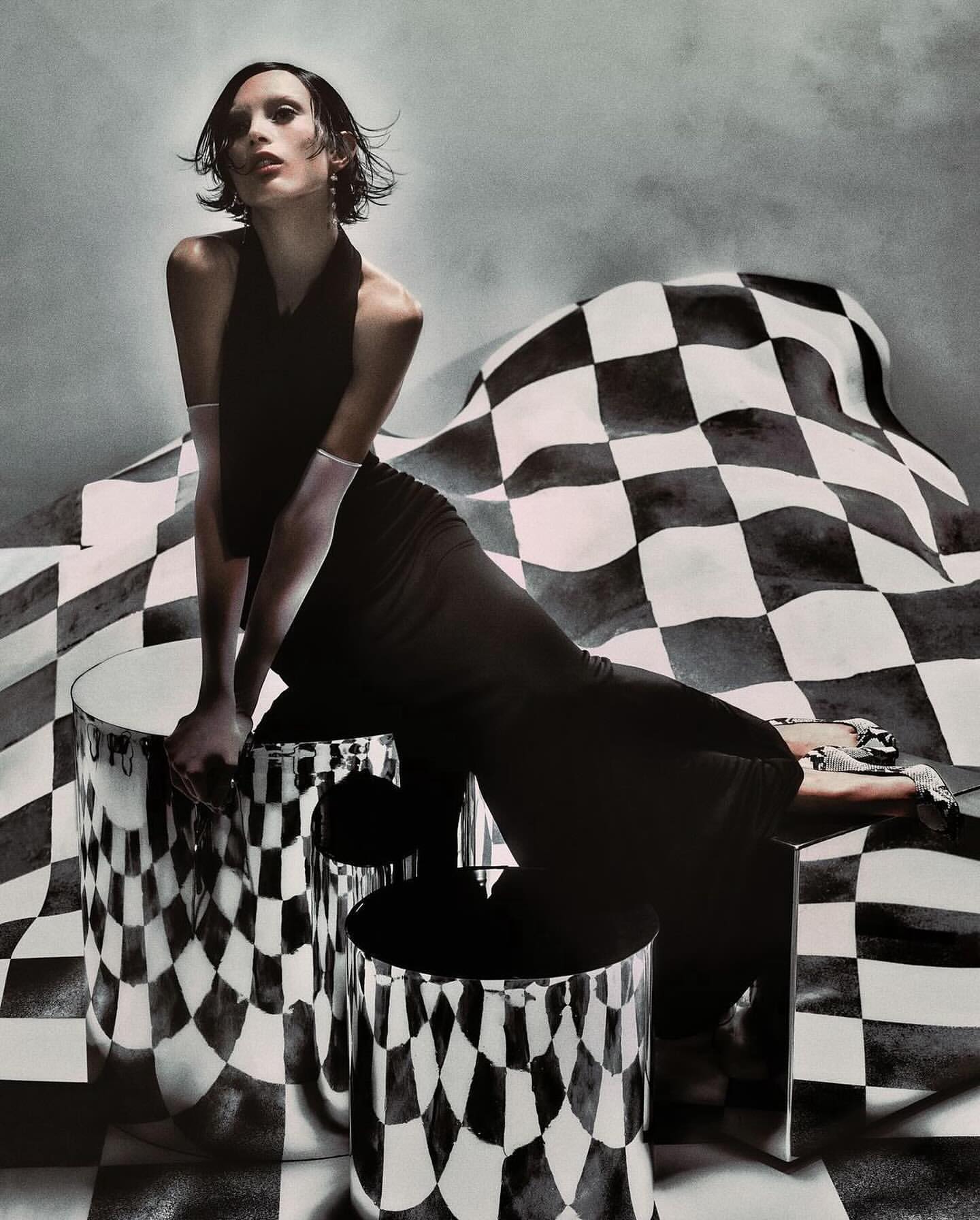 An adult female model poses in a black dress, seated on metallic cylindrical objects, with a checkered backdrop for a fashion editorial.