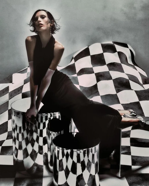 An adult female model poses in a black dress, seated on metallic cylindrical objects, with a checkered backdrop for a fashion editorial.