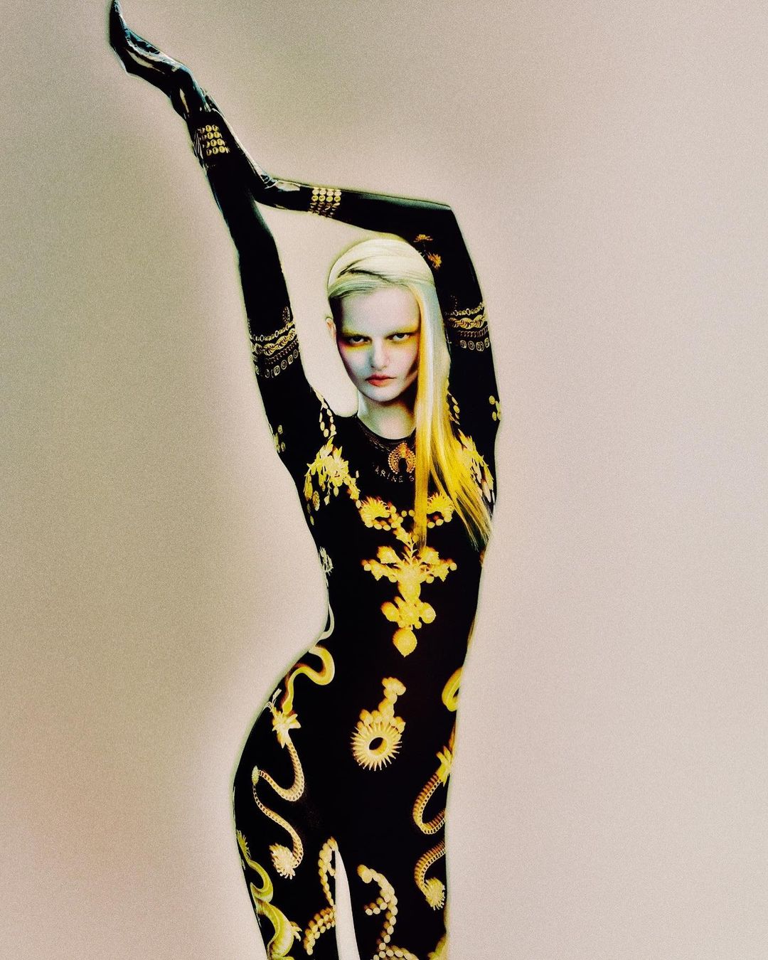 A woman with blond hair poses in a black and yellow patterned bodysuit against a neutral background, arms raised above her head.