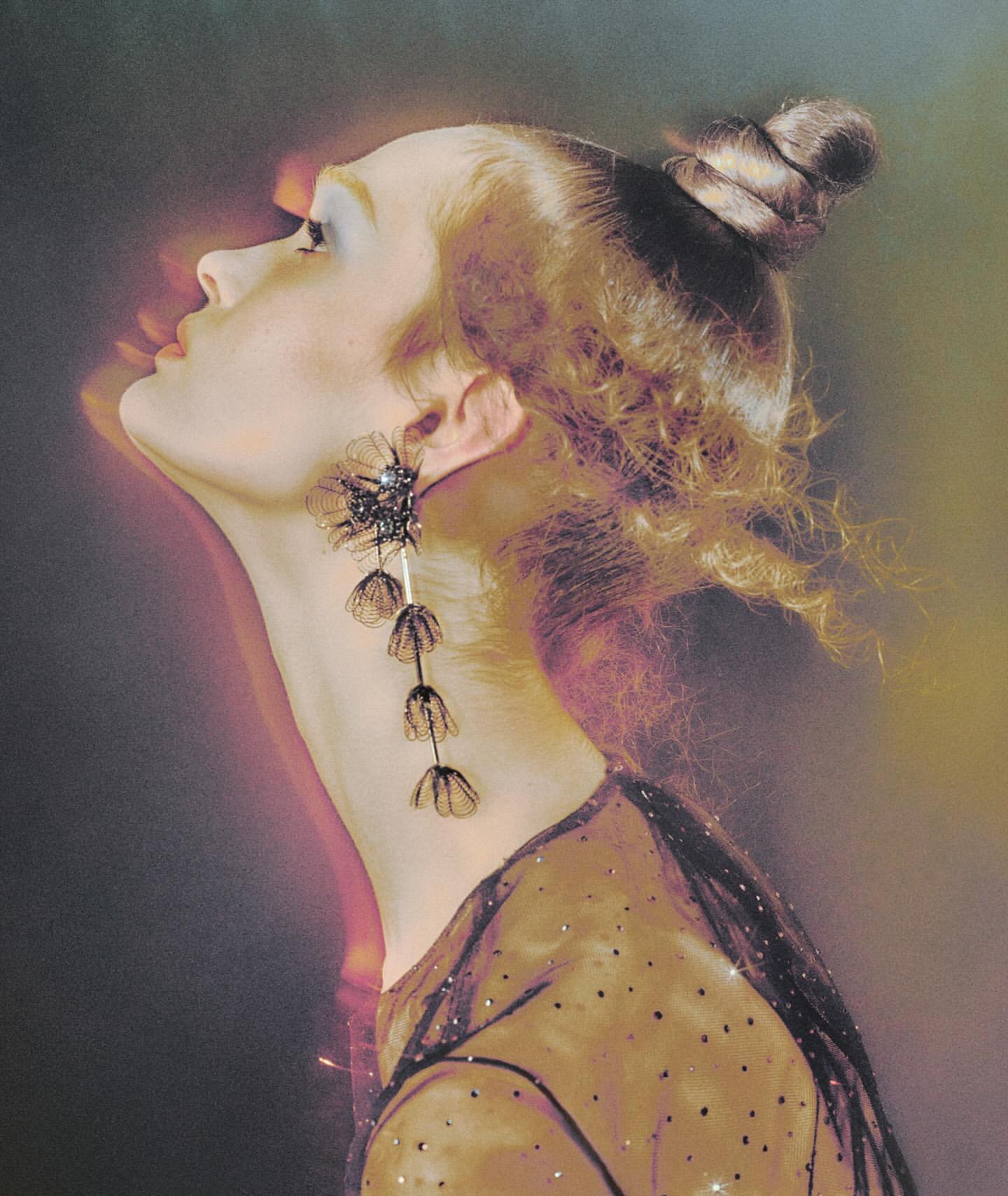 Profile shot of a woman with auburn hair in an updo, wearing a sheer, sparkly top and large floral earrings, looking upward.