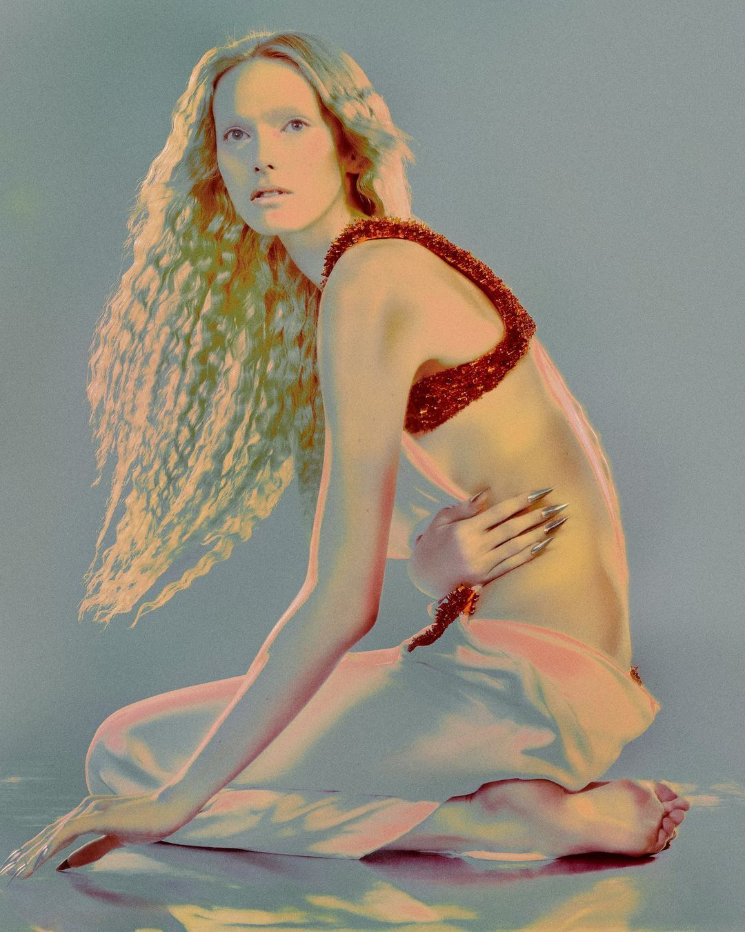 Woman with long blonde curly hair poses in front of a soft gradient background wearing a beaded top and pale skirt in a studio shot.