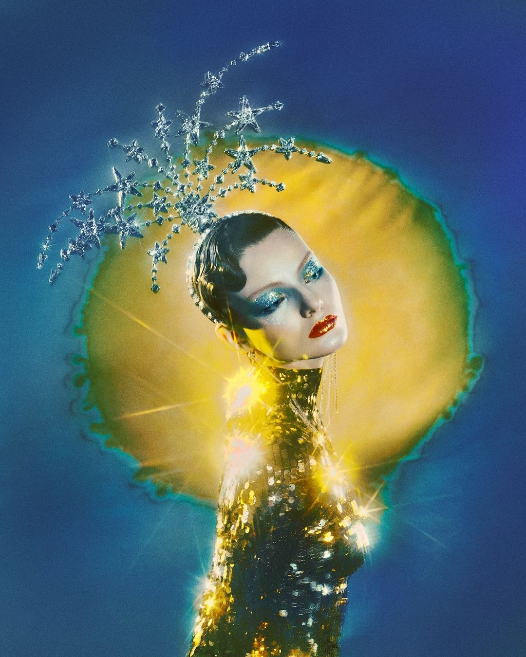 Woman with decorative jeweled star headpiece and dramatic eye makeup poses in gold sequin dress against a soft blue and gold background.