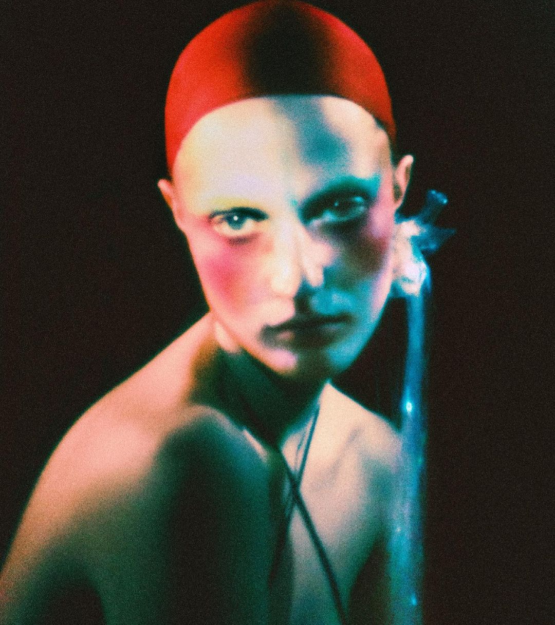 Striking portrait of a bald adult with a red cap, accentuated by pink and blue lighting against a dark backdrop. The shot is intimate and intense.