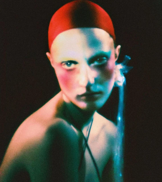 Striking portrait of a bald adult with a red cap, accentuated by pink and blue lighting against a dark backdrop. The shot is intimate and intense.