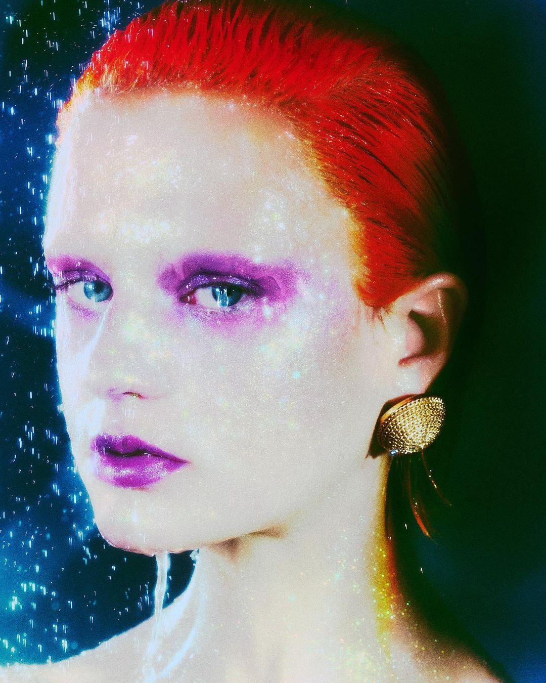 Close-up portrait of a woman with red hair and artistic purple makeup, posing against a dark, out-of-focus background.