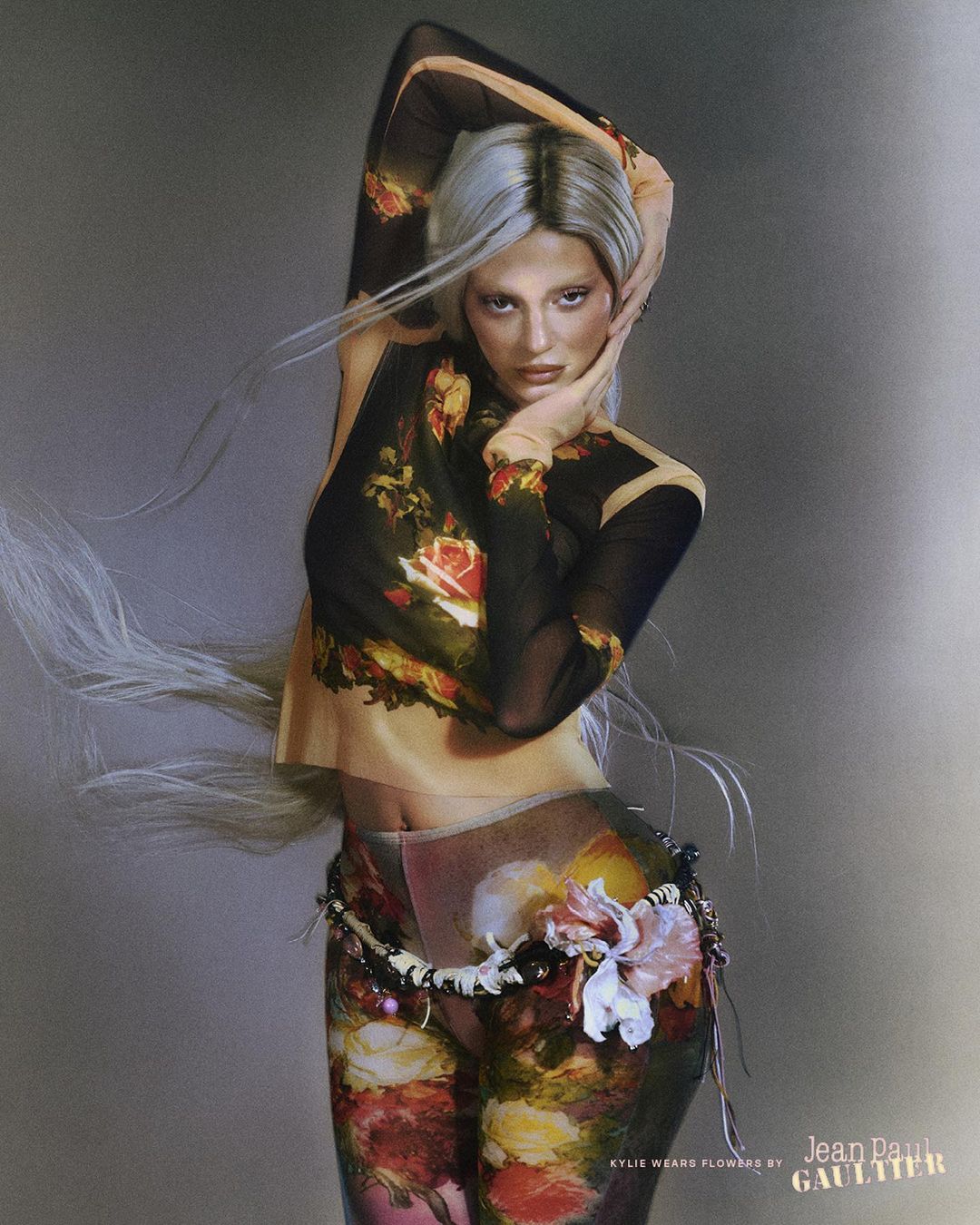 Female fashion model with silver hair poses in a sheer floral top and pants with a decorative belt against a grey background.