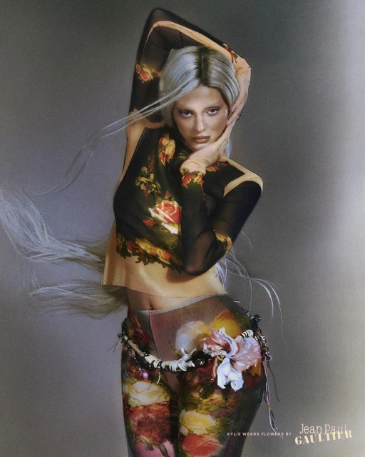 Female fashion model with silver hair poses in a sheer floral top and pants with a decorative belt against a grey background.