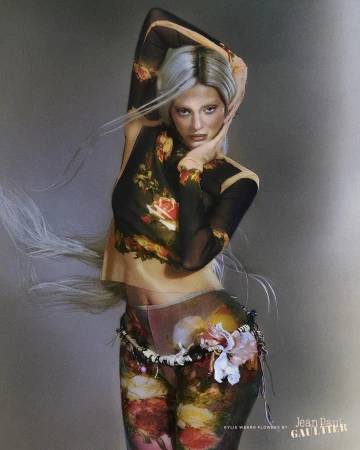 Female fashion model with silver hair poses in a sheer floral top and pants with a decorative belt against a grey background.