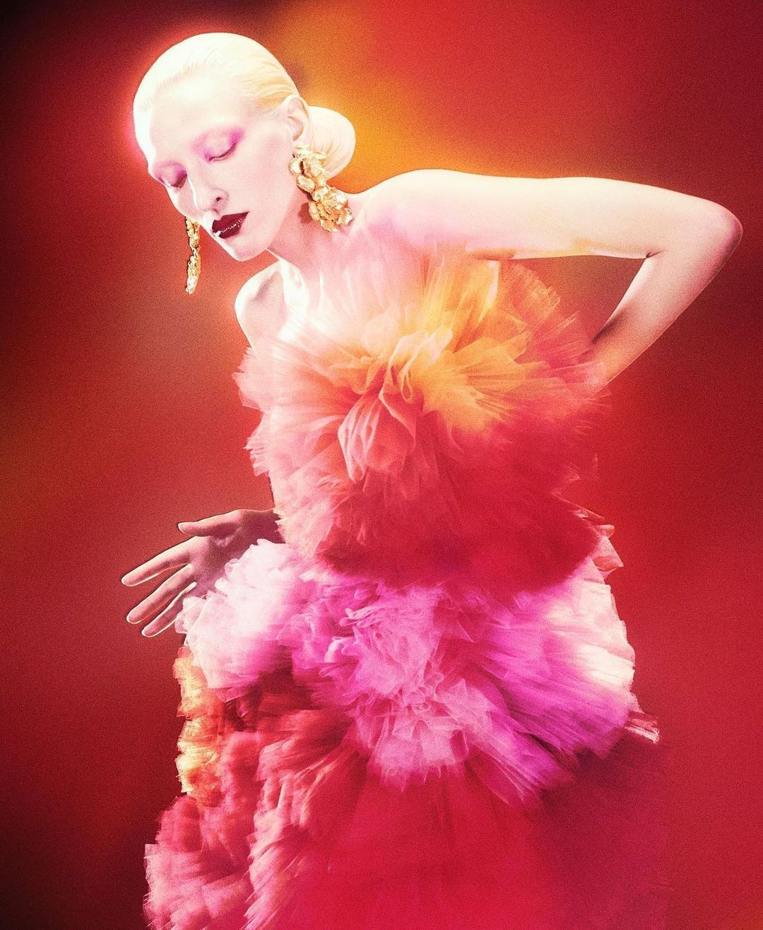 Woman with pale skin and blonde hair poses in pink and orange ruffled gown against a red background, wearing large gold earrings.