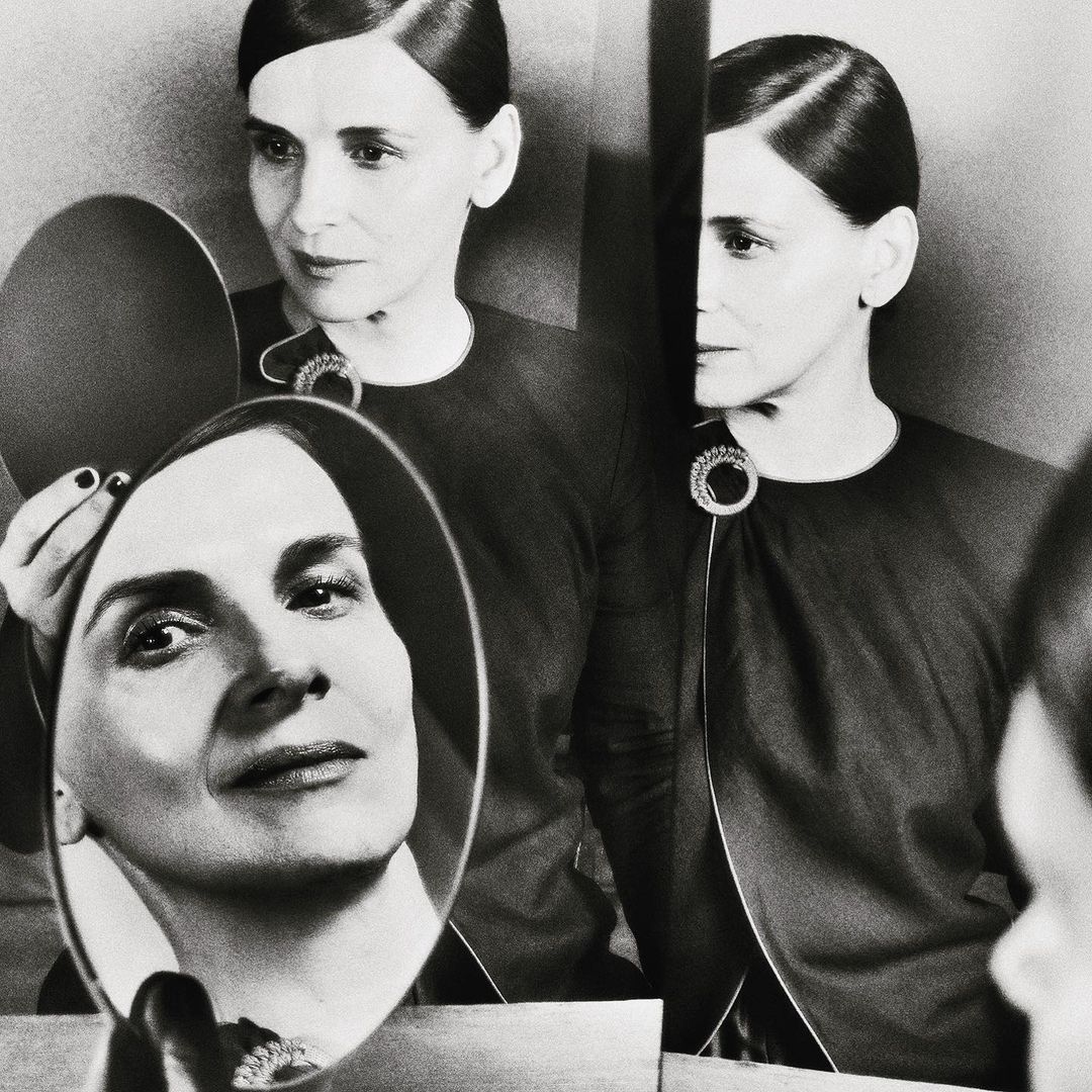 Black-and-white portrait of an elegant mature woman with dark hair and an embellished dark dress, reflected in a hand mirror.