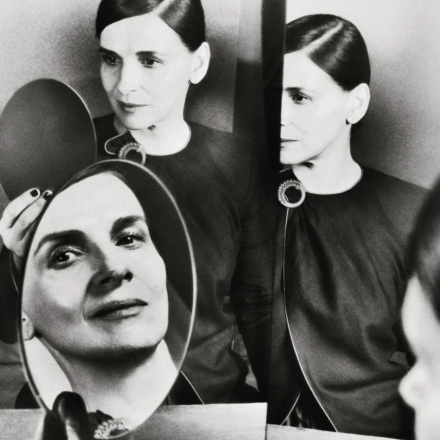 Black-and-white portrait of an elegant mature woman with dark hair and an embellished dark dress, reflected in a hand mirror.