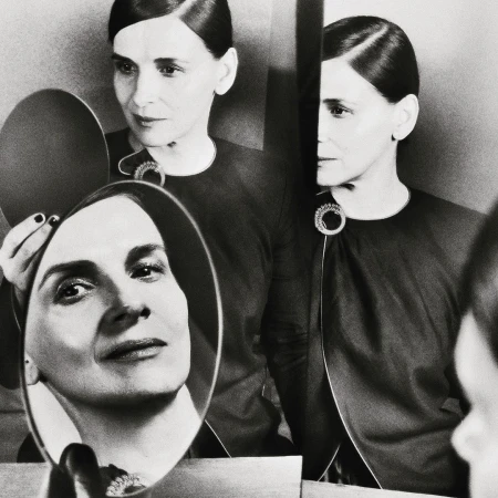 Black-and-white portrait of an elegant mature woman with dark hair and an embellished dark dress, reflected in a hand mirror.