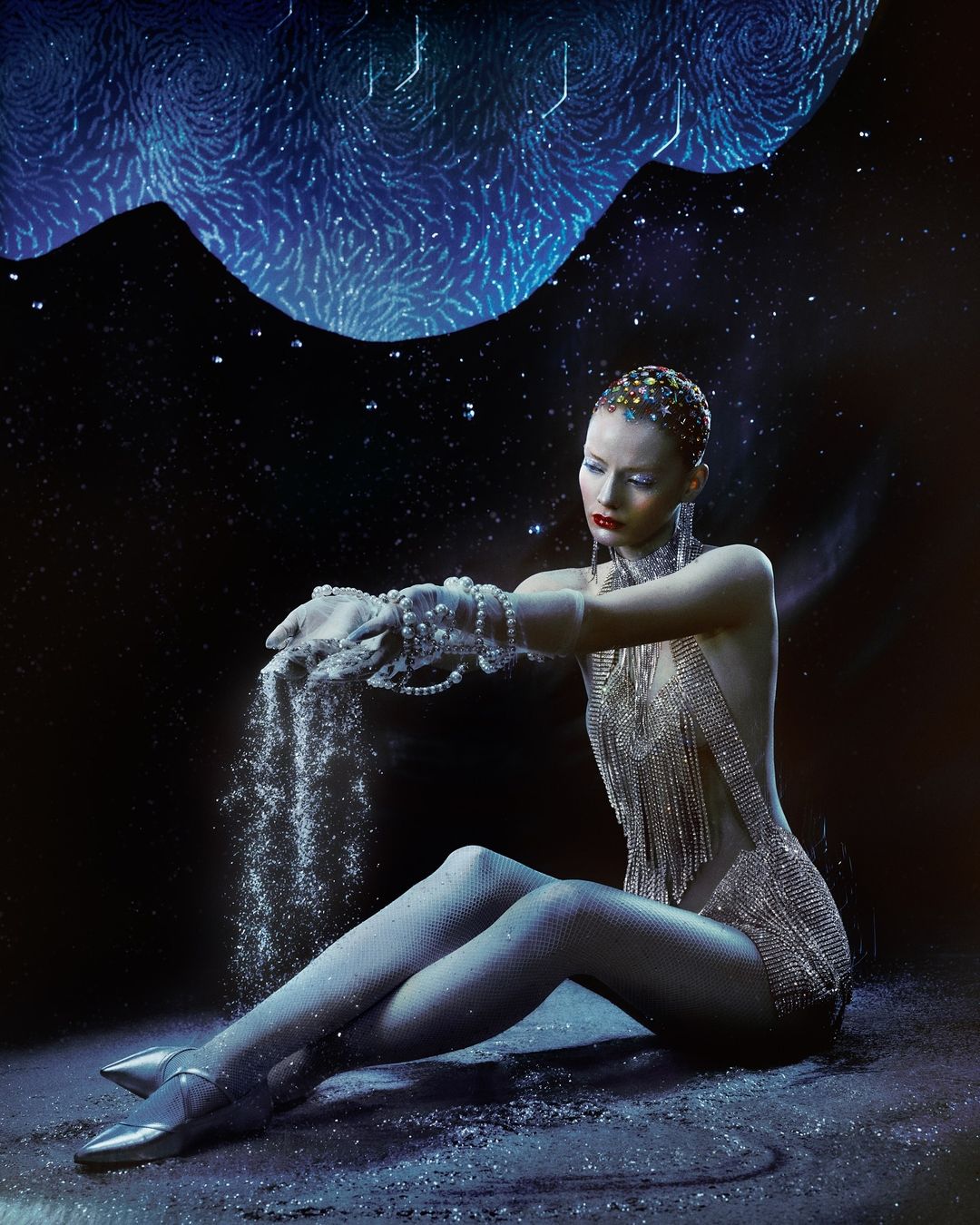 Woman in glittering bodysuit sits holding shimmering powder against a backdrop of swirling blue lights, shot in a photo studio.