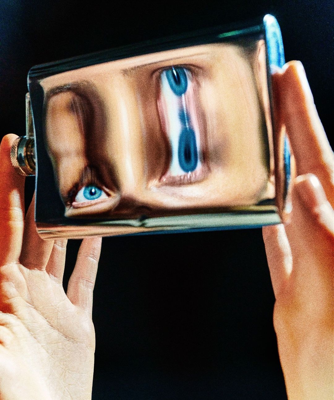 Distorted face reflection in a stainless steel flask held by two hands against a dark background, creating an abstract visual.