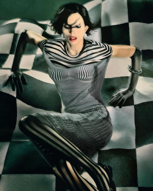 Fashion shot of a woman with dark hair, wearing a striped outfit, posing on a black and white checkerboard surface in a studio.