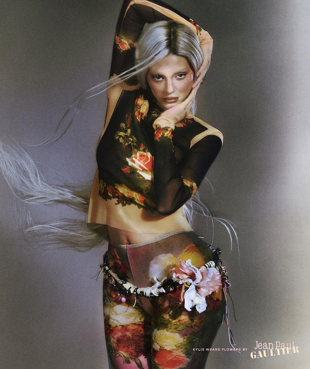 A woman with silver hair poses in a sheer, floral-print top and leggings in a studio. Jean Paul Gaultier logo visible at bottom right.