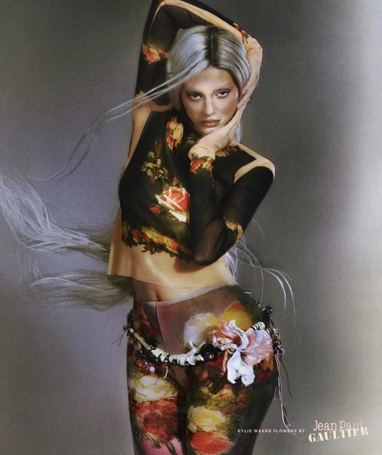 A woman with silver hair poses in a sheer, floral-print top and leggings in a studio. Jean Paul Gaultier logo visible at bottom right.