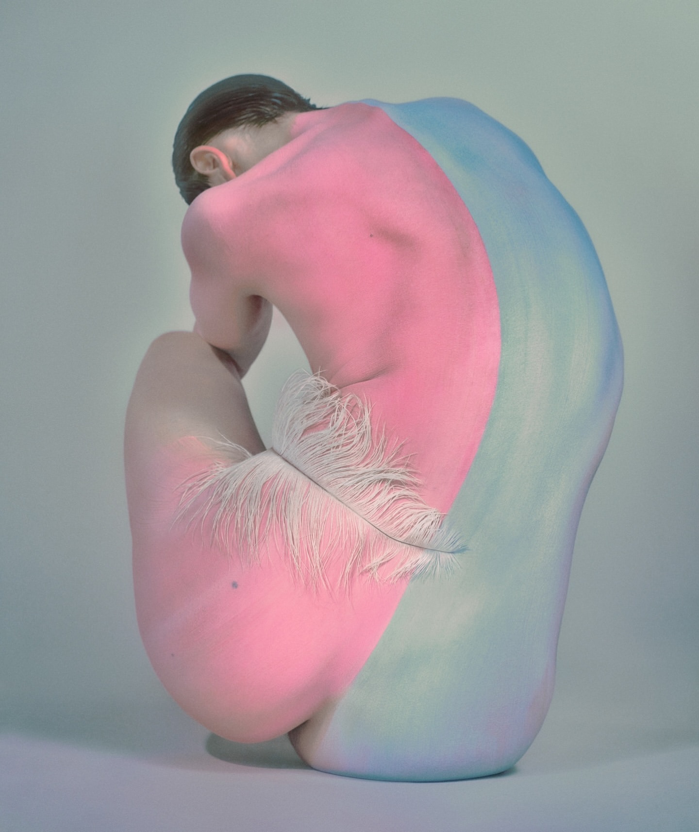 Artistic nude crouched figure painted in pink and blue with a feather. Conceptual image on a light background with soft lighting.