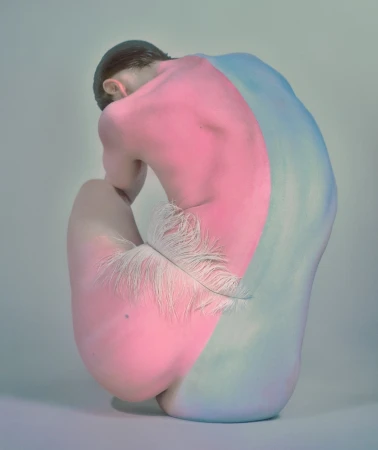 Artistic nude crouched figure painted in pink and blue with a feather. Conceptual image on a light background with soft lighting.