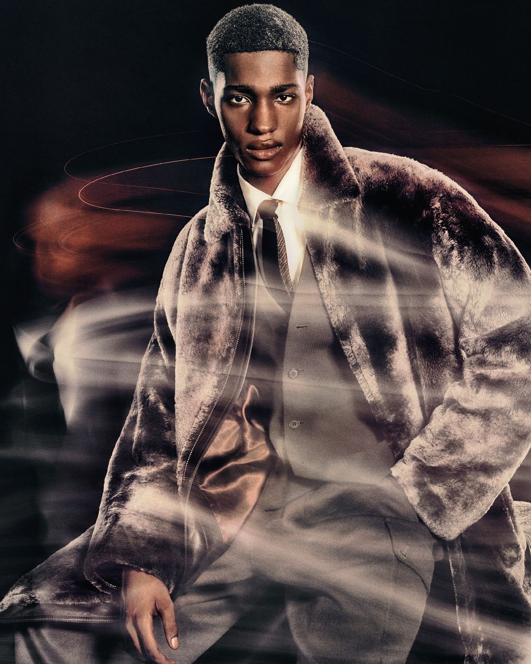 Portrait of a young adult male fashion model in a faux fur coat, standing indoors against a dark background, with long exposure light streaks.