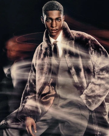 Portrait of a young adult male fashion model in a faux fur coat, standing indoors against a dark background, with long exposure light streaks.