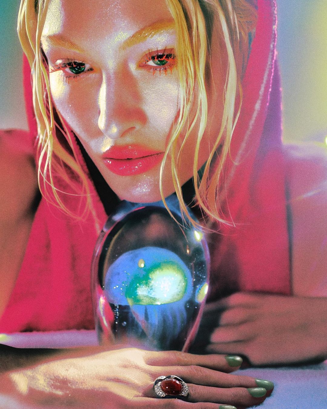 Close-up shot of a fair-skinned adult female model with blonde hair, pink hooded sweatshirt, and large ring holding a glass orb.