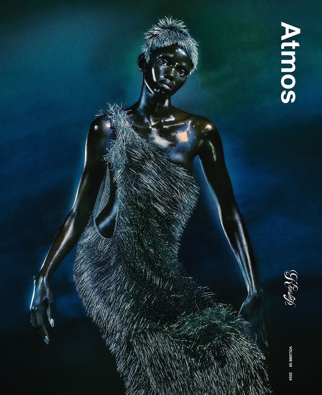 A model with short, spiky hair and dark skin poses in a fringed dress against a muted teal and blue background for Atmos magazine.