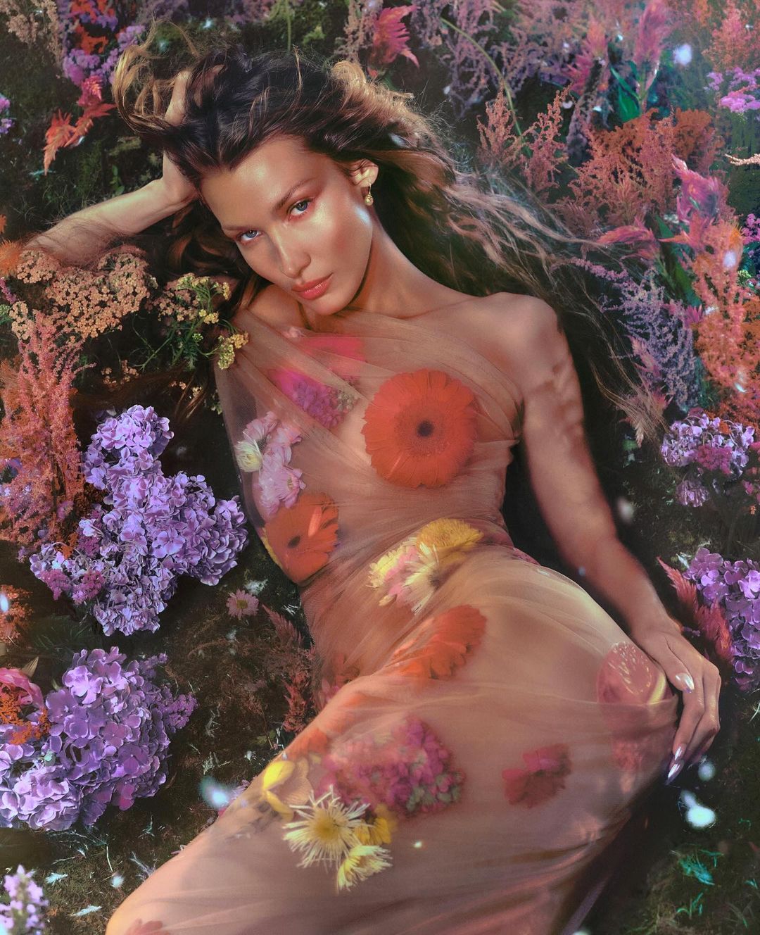 A model poses in a field of flowers, draped in a sheer garment with colorful flowers, capturing a blend of beauty and nature.