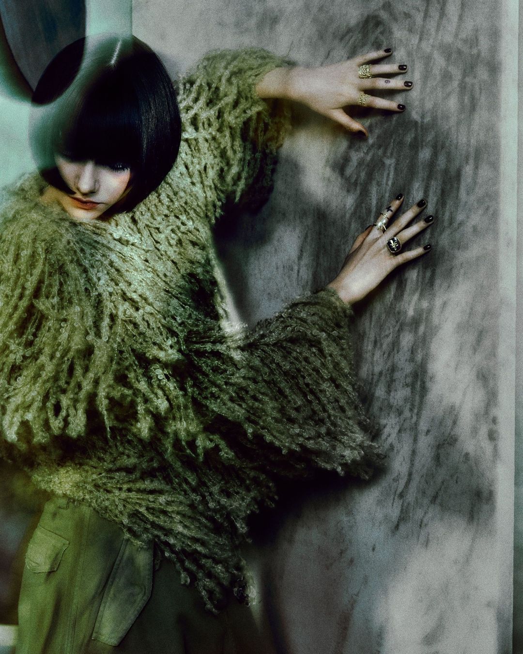 Fashion photo of a woman in a fringed top and green pants leaning against a mottled wall, showcasing unique textures and a moody aesthetic.