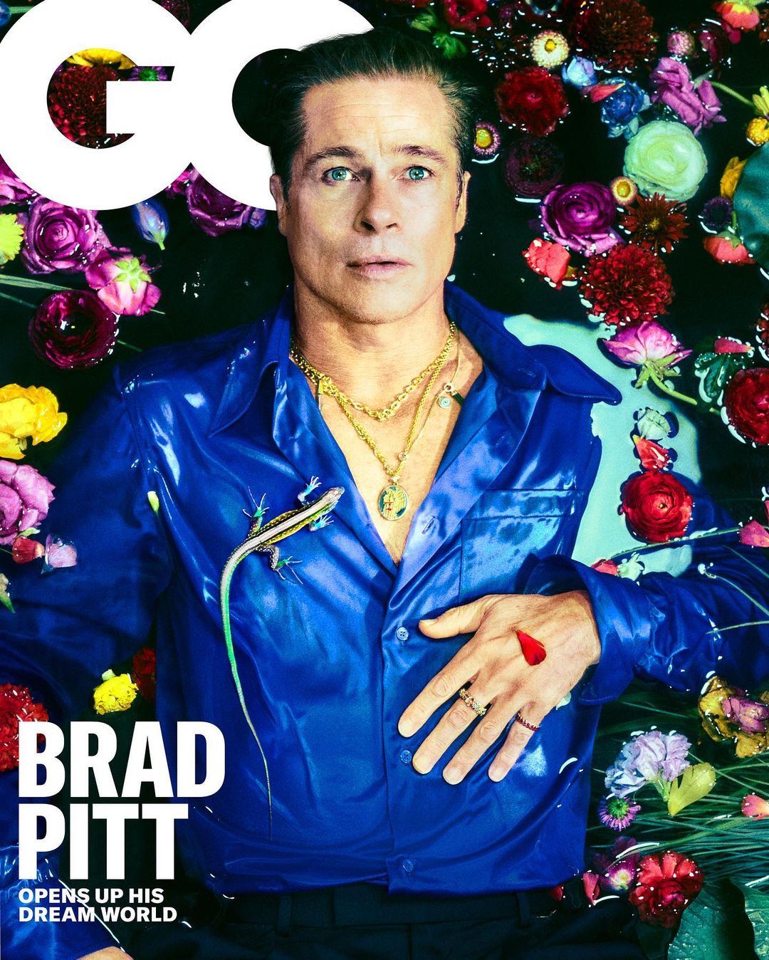 Brad Pitt is captured in a close-up portrait, wearing a shiny blue shirt and gold necklaces while lying amidst flowers and water.