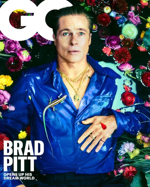 Brad Pitt is captured in a close-up portrait, wearing a shiny blue shirt and gold necklaces while lying amidst flowers and water.