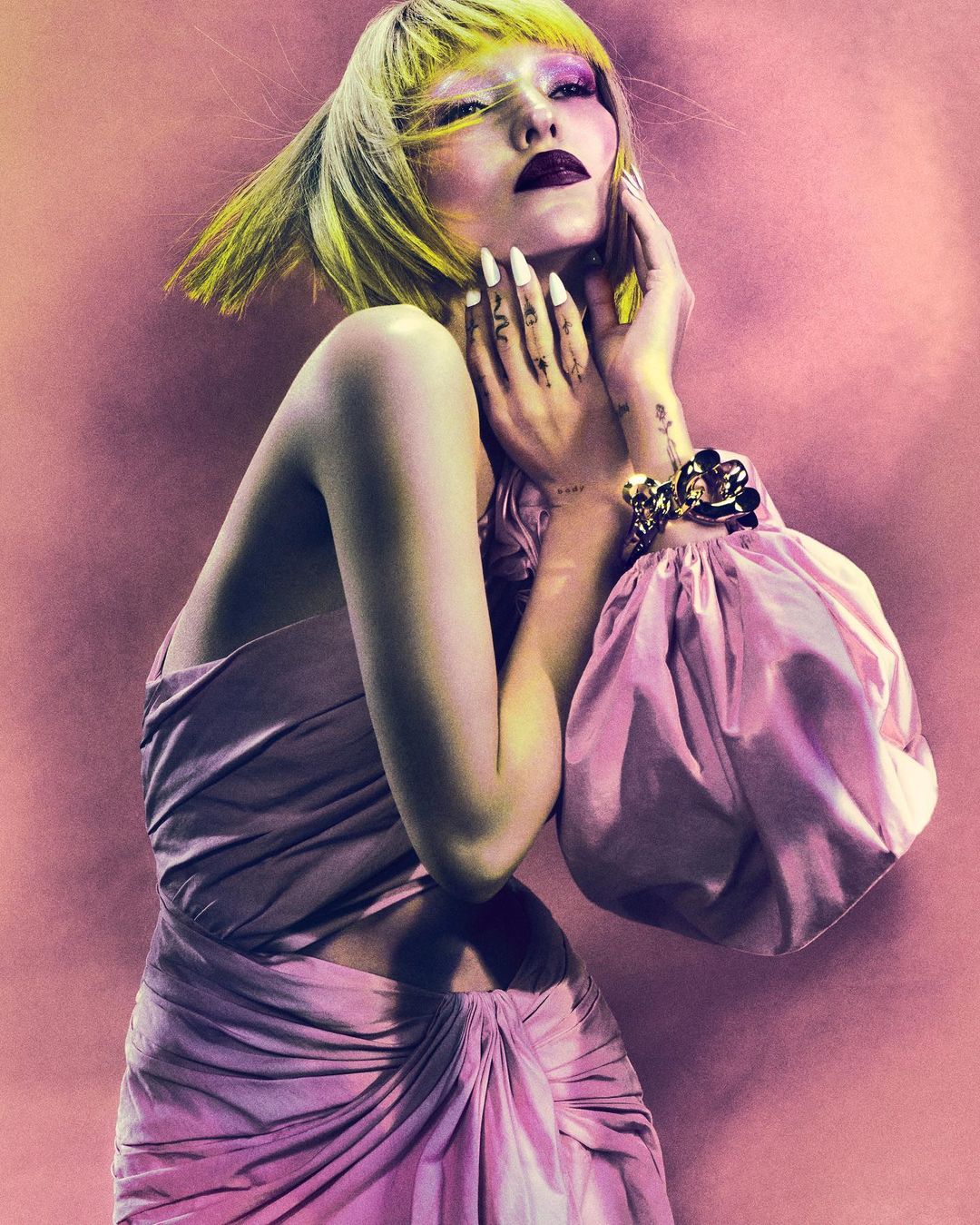 Woman with yellow hair, heavy makeup, and pink gown poses against pink backdrop with hands near face, wearing large gold bracelet.
