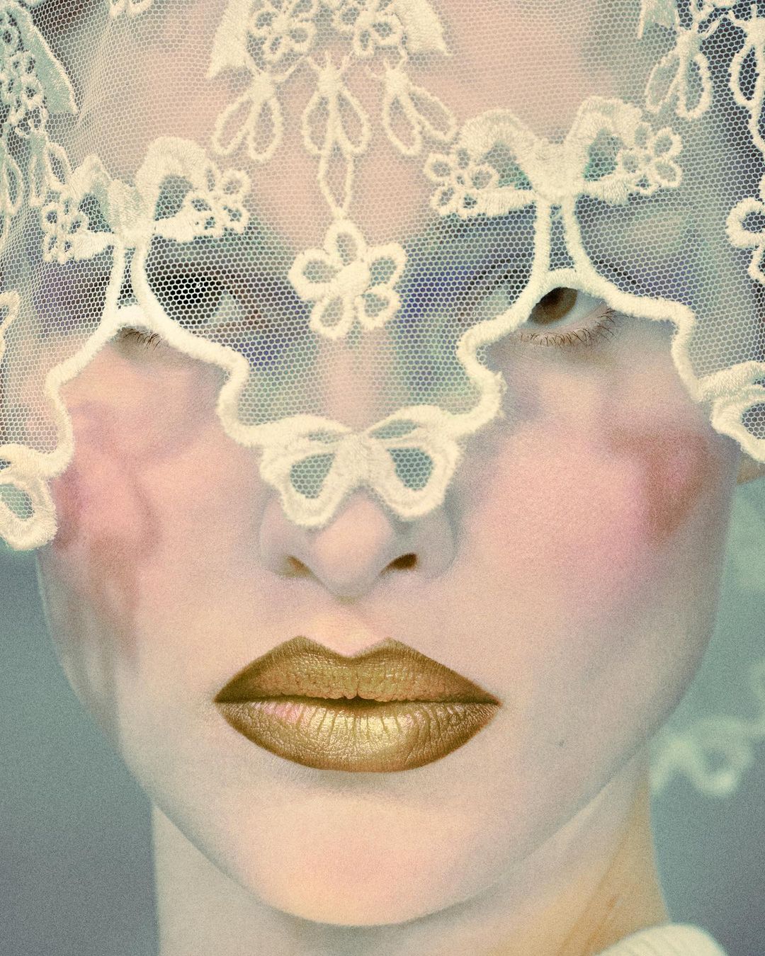 Close-up of a person with a pale complexion and gold lipstick, wearing a lacy white veil that partially obscures their face in a portrait shot.