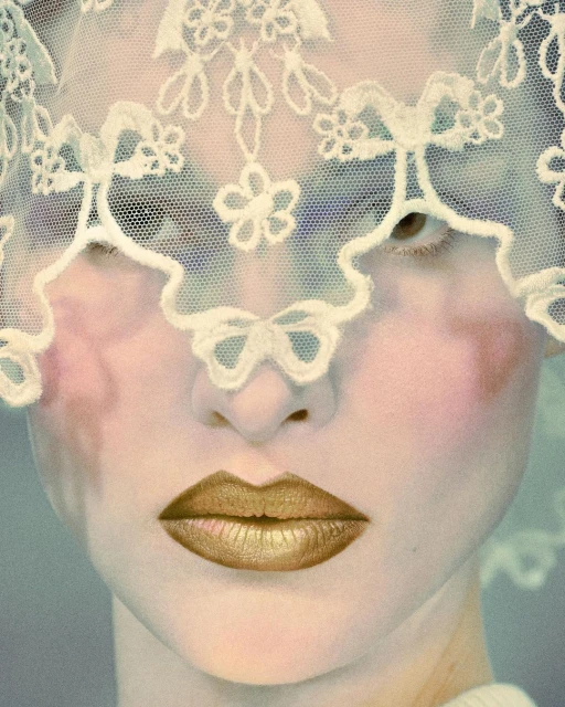Close-up of a person with a pale complexion and gold lipstick, wearing a lacy white veil that partially obscures their face in a portrait shot.