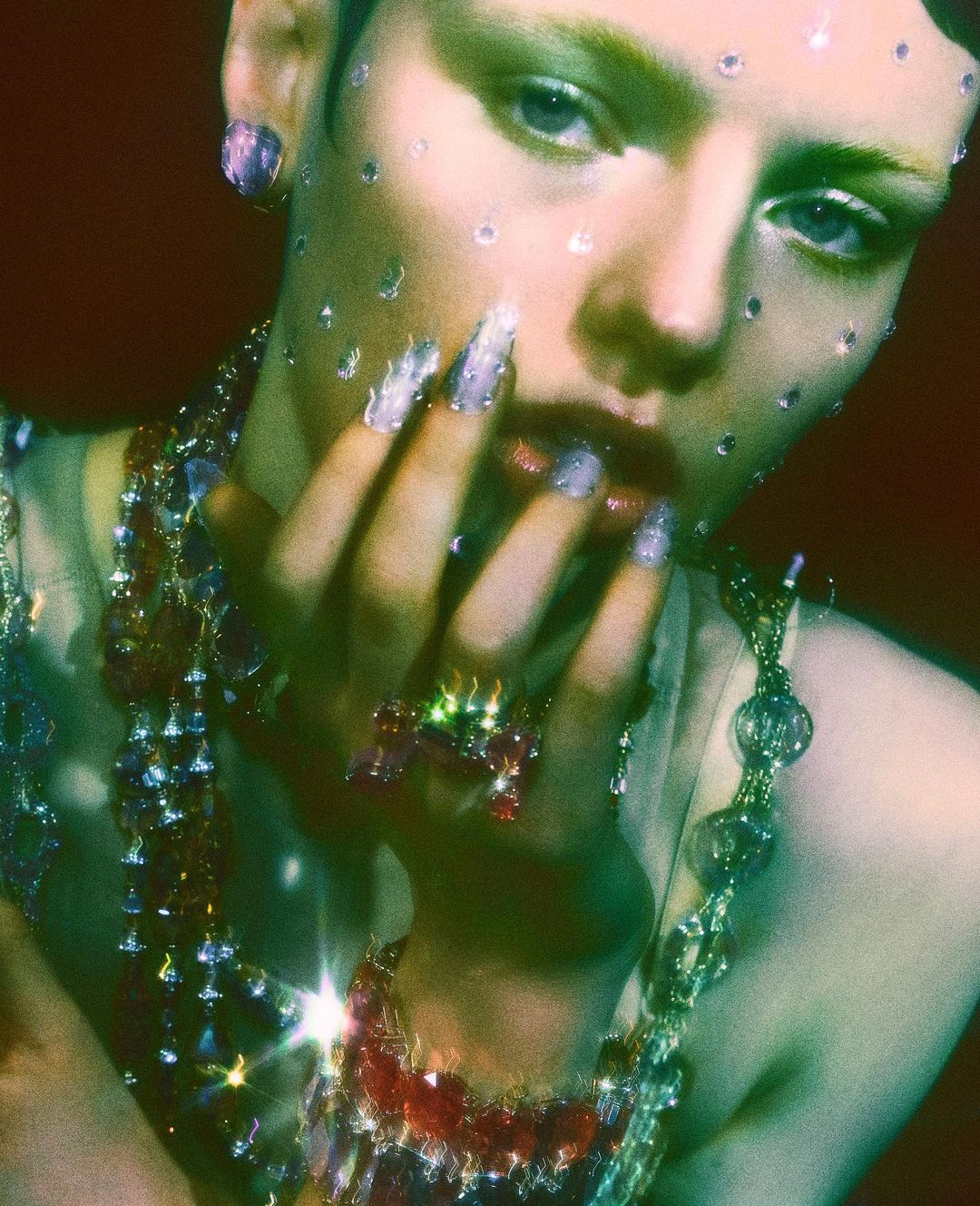 Close-up of a fair-skinned adult female with fair hair and green eyeshadow adorned with jewelry and small transparent gems on her face.