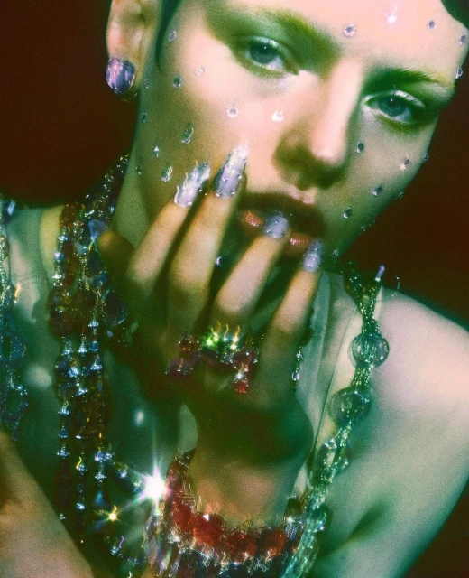 Close-up of a fair-skinned adult female with fair hair and green eyeshadow adorned with jewelry and small transparent gems on her face.
