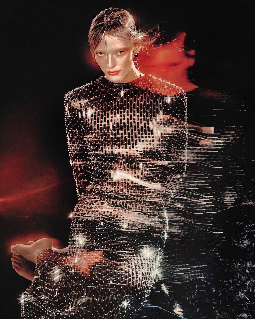 A fashionable woman with red lipstick and fair skin poses in a shiny, beaded long-sleeve dress against a dark background in a studio.