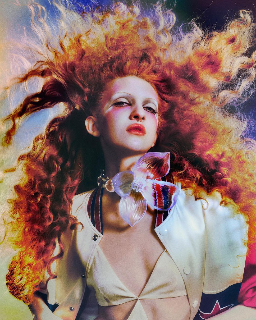 Young woman with elaborate hair and makeup wearing a large floral necklace and jacket, posing against a colorful, blurred background.