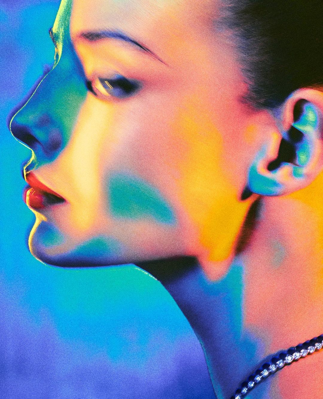 Close-up portrait of a woman's profile with vibrant blue, yellow, and pink lighting, highlighting her face and diamond necklace.
