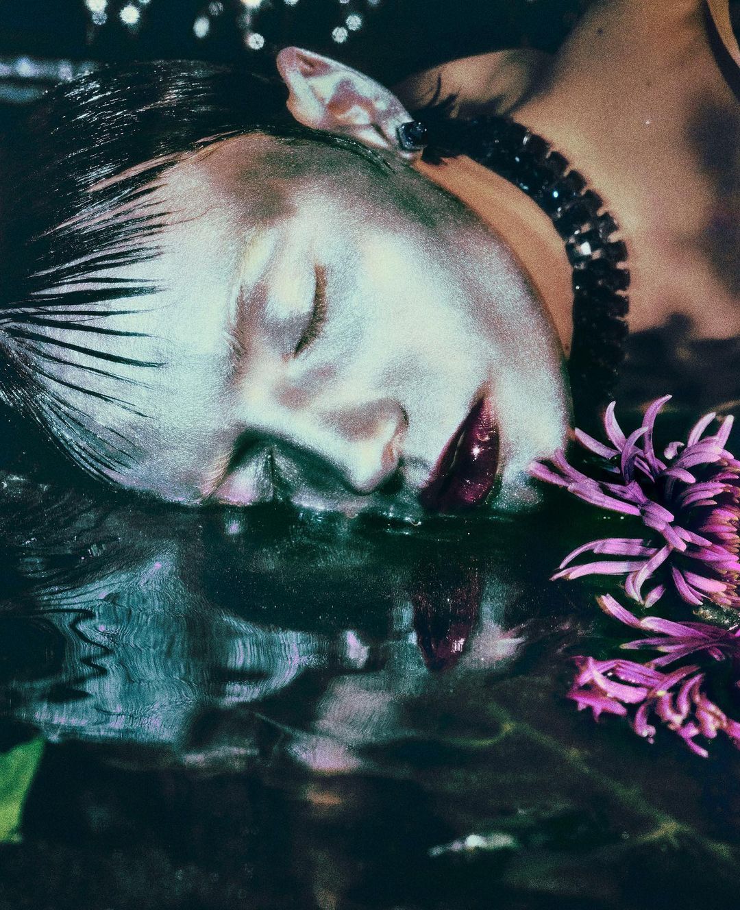 Close-up of a fair-skinned woman with closed eyes and dark lipstick, adorned with a beaded choker and flower, lying in water.