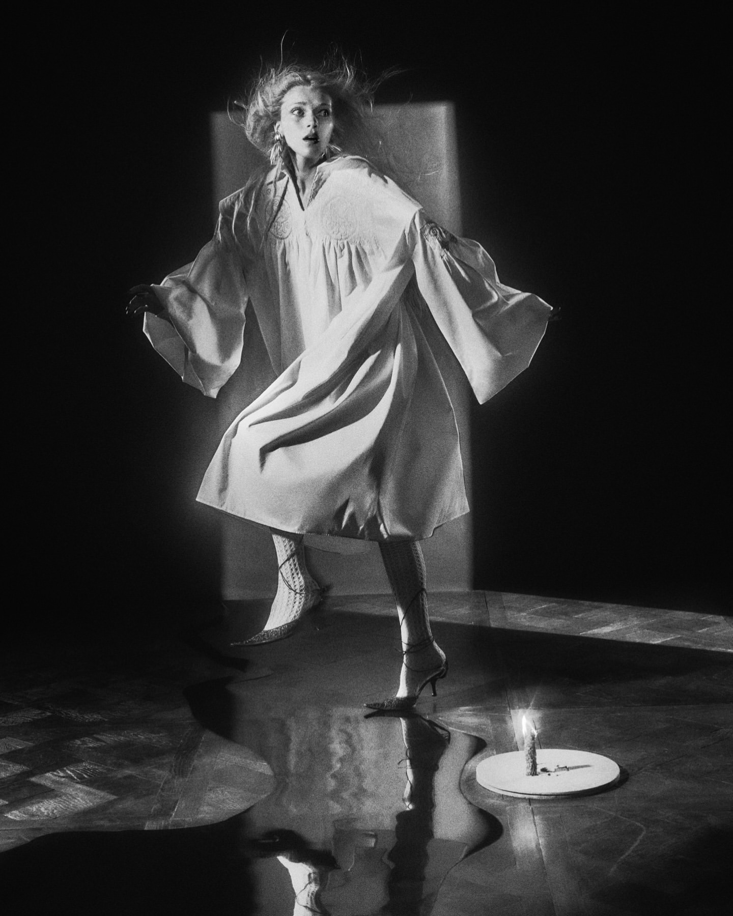 Monochrome fashion shot: woman in robe and heels strikes dramatic pose near candle, spotlit against a dark background.