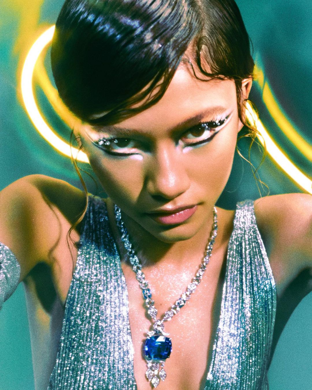 Close-up of a woman with elaborate eye makeup and a large blue gem pendant necklace against a soft green and yellow light background.