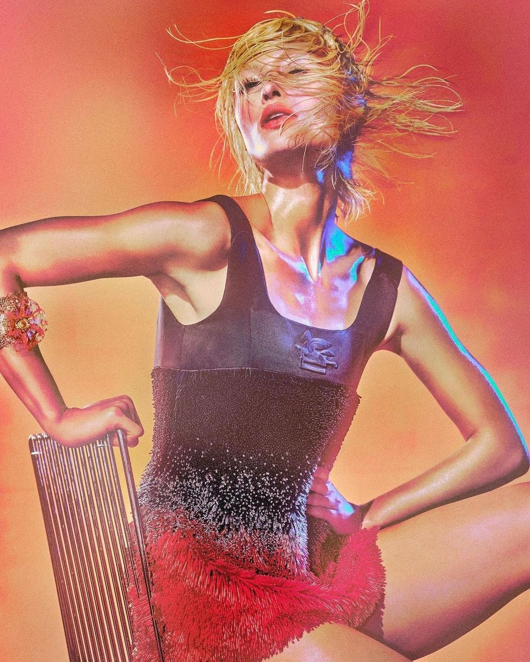 Woman with wet blond hair poses while seated in a black and red beaded outfit in a studio environment with vibrant, colorful lighting.
