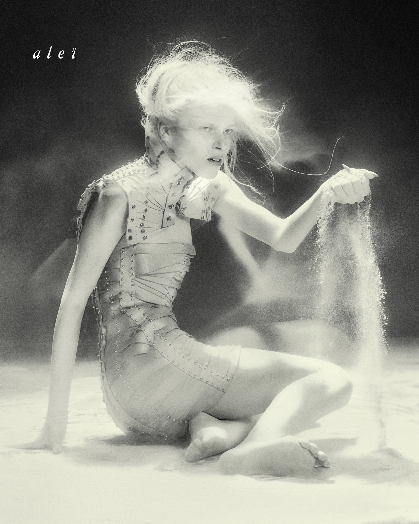 Monochrome portrait of a woman with bright hair kneeling in what appears to be sand, lightly holding grains falling through her fingers.