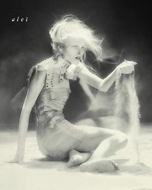 Monochrome portrait of a woman with bright hair kneeling in what appears to be sand, lightly holding grains falling through her fingers.