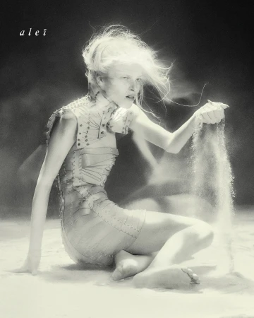 Monochrome portrait of a woman with bright hair kneeling in what appears to be sand, lightly holding grains falling through her fingers.