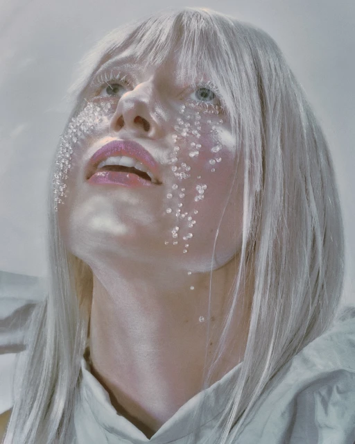 Portrait of a woman with platinum blonde hair and striking makeup, featuring jewel-like accents on her face, gazing upwards in a serene setting.