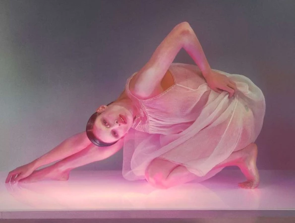 Woman in a sheer white dress kneeling and bending backward in vibrant pink lighting against a neutral background.