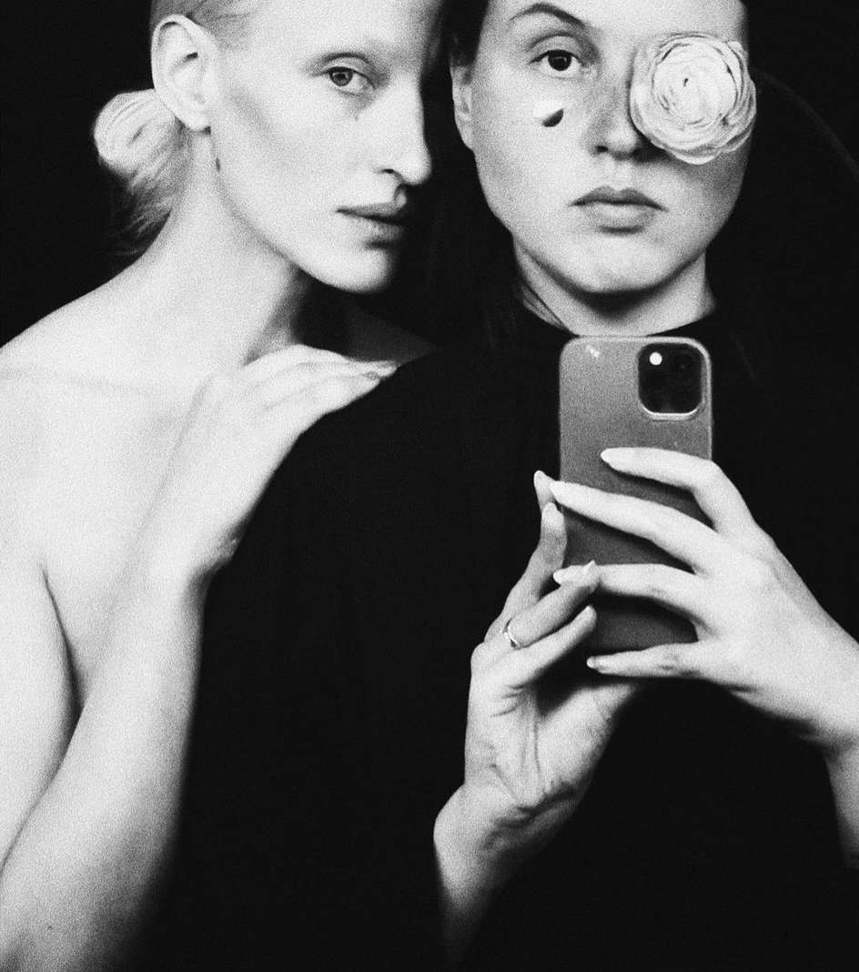 Monochrome portrait of two fair-skinned adult women, one holding a smartphone for a selfie, and a white ranunculus flower over one eye.